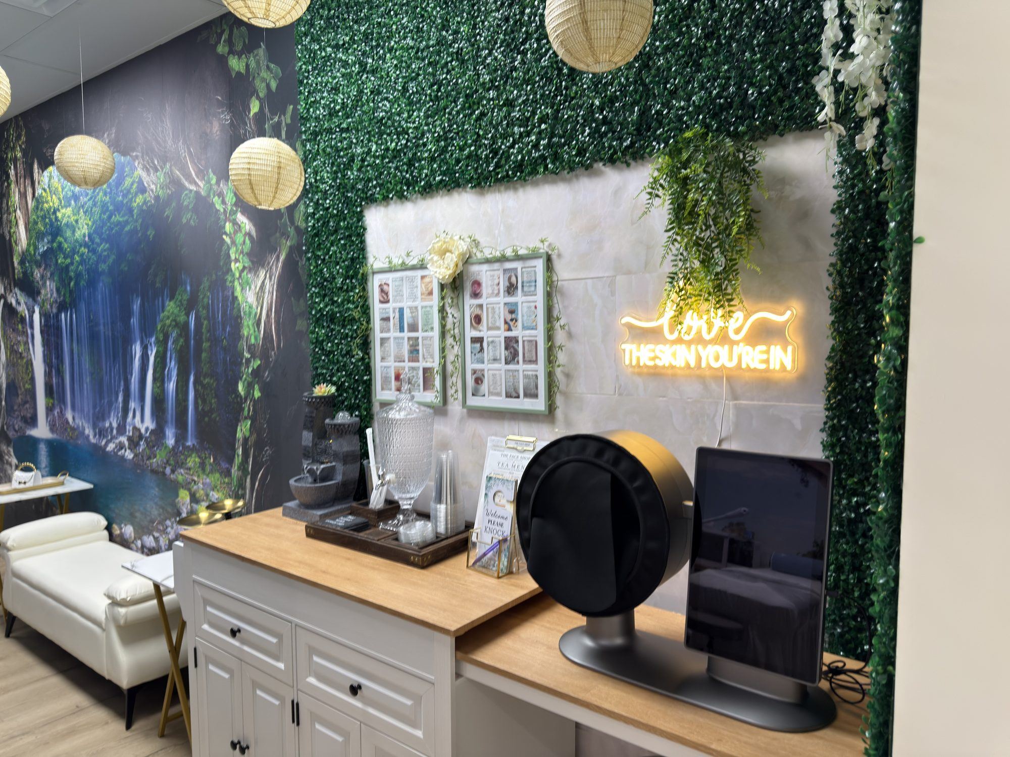 Spa reception with a green foliage wall, waterfall mural, paper lanterns and a 'Love Yourself' neon sign